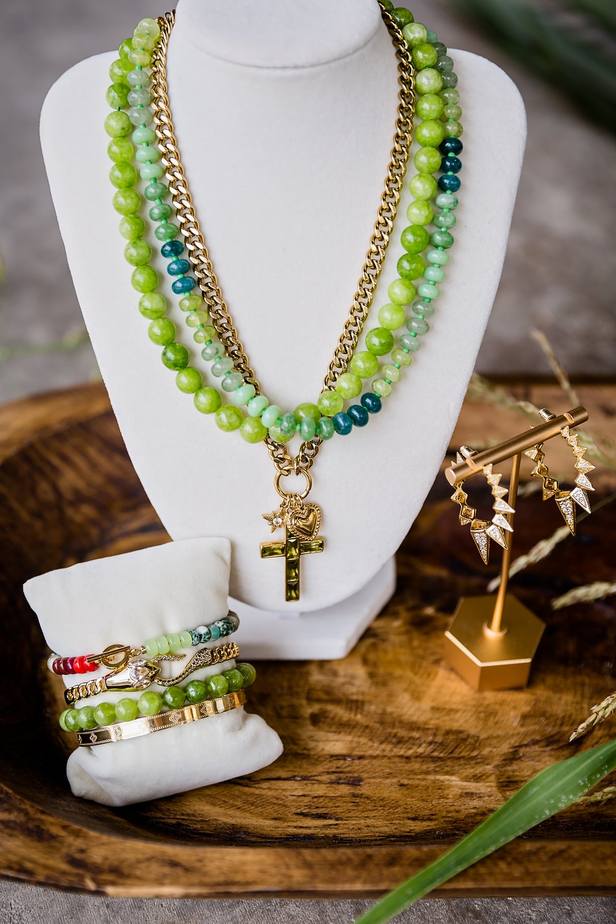 A layered necklace set displayed on a white mannequin bust, featuring green and teal beaded strands with gold-tone chains and a cross pendant, alongside matching beaded bracelets on a small white stand. The setup is styled on a wooden tray with decorative dried grass and jewelry stands.