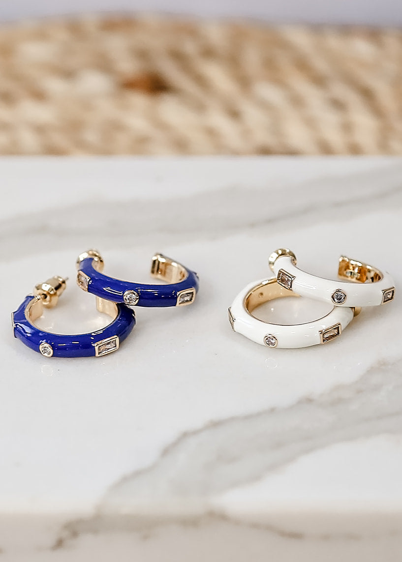A pair of enamel hoop earrings with gold plating and small cubic zirconia accents, placed on a marble surface with a woven mat behind.