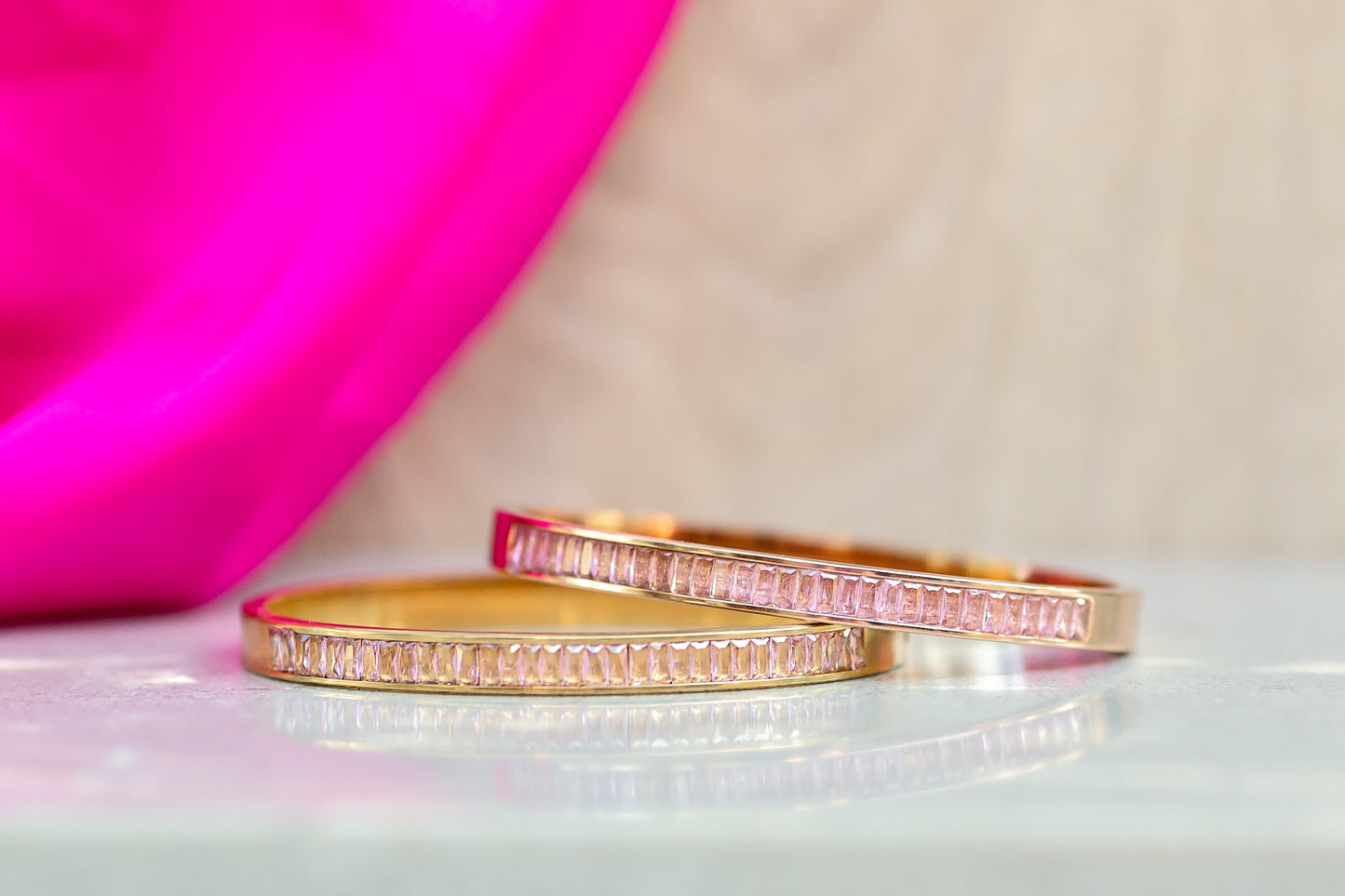 Two gold bracelets with pink accents on a light pink surface.