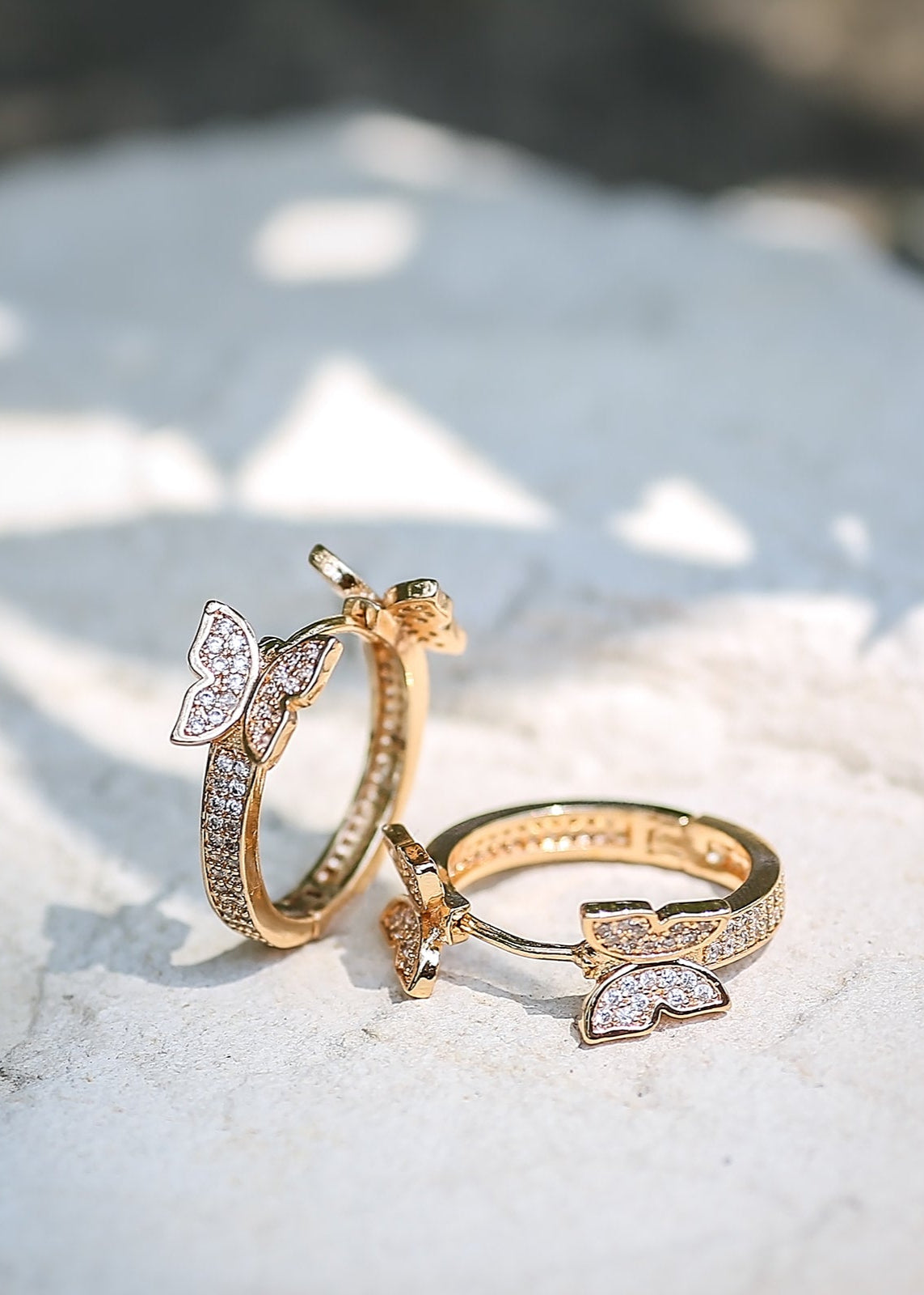 A pair of gold-colored hoop earrings with butterfly designs and micro pavé cubic zirconia stones, placed on a white textured surface.