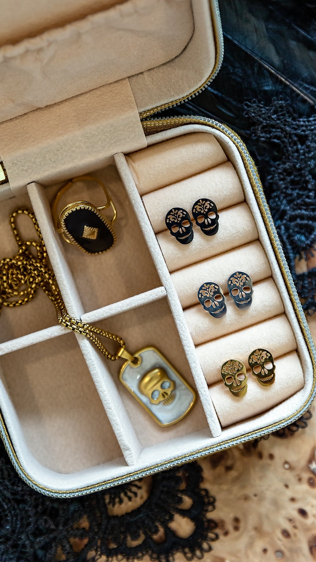An open jewelry organizer case with cream velvet compartments holding a gold pendant necklace, a black-and-gold ring, and multiple pairs of skull stud earrings in black, blue, and gold.