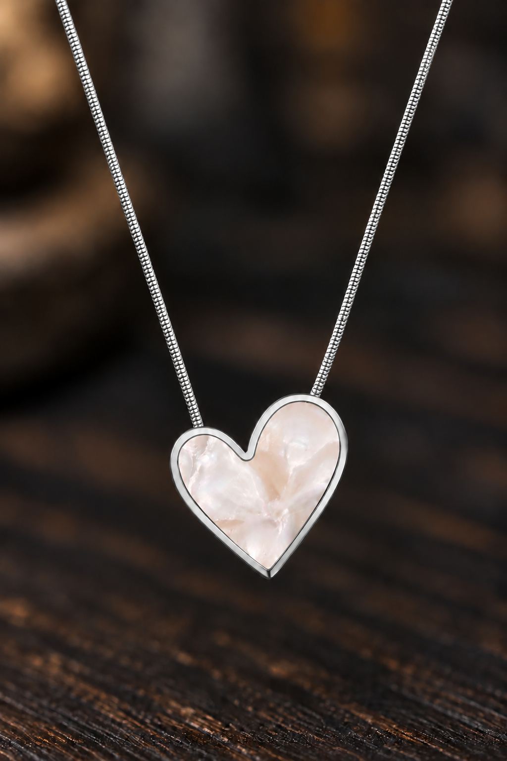 A silver heart pendant necklace with a pale pink mother-of-pearl center, suspended on a delicate silver chain against a dark wooden background.
