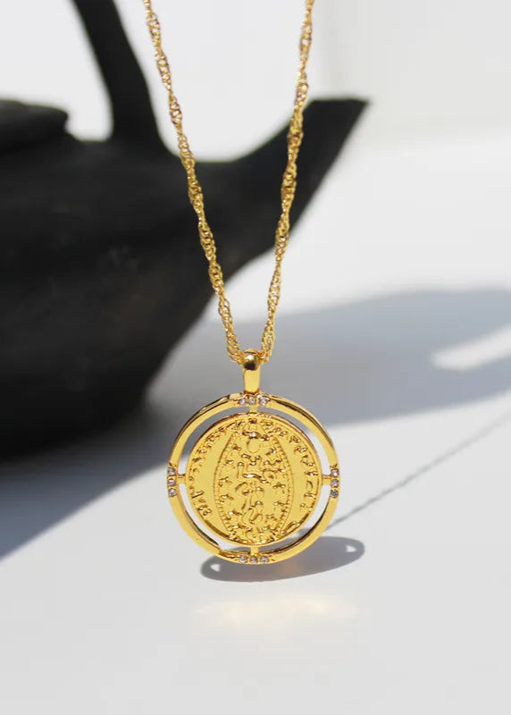A gold-plated necklace with intricate chain design and an embossed charm detail, displayed on a white background next to a black object, likely a bird or a sculpture.