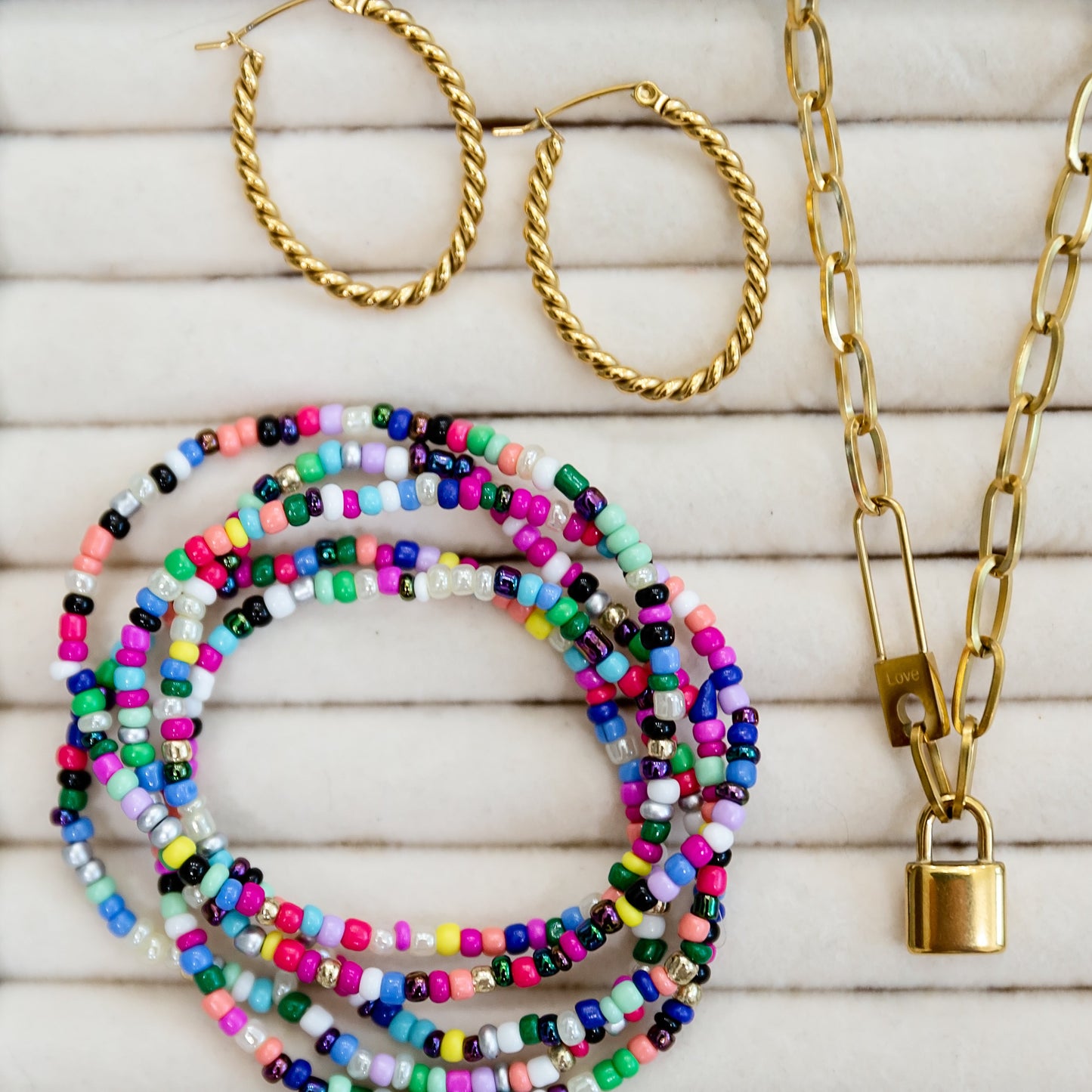 Gold hoop earrings, colorful beaded bracelets, and a gold chain necklace on a wooden surface.