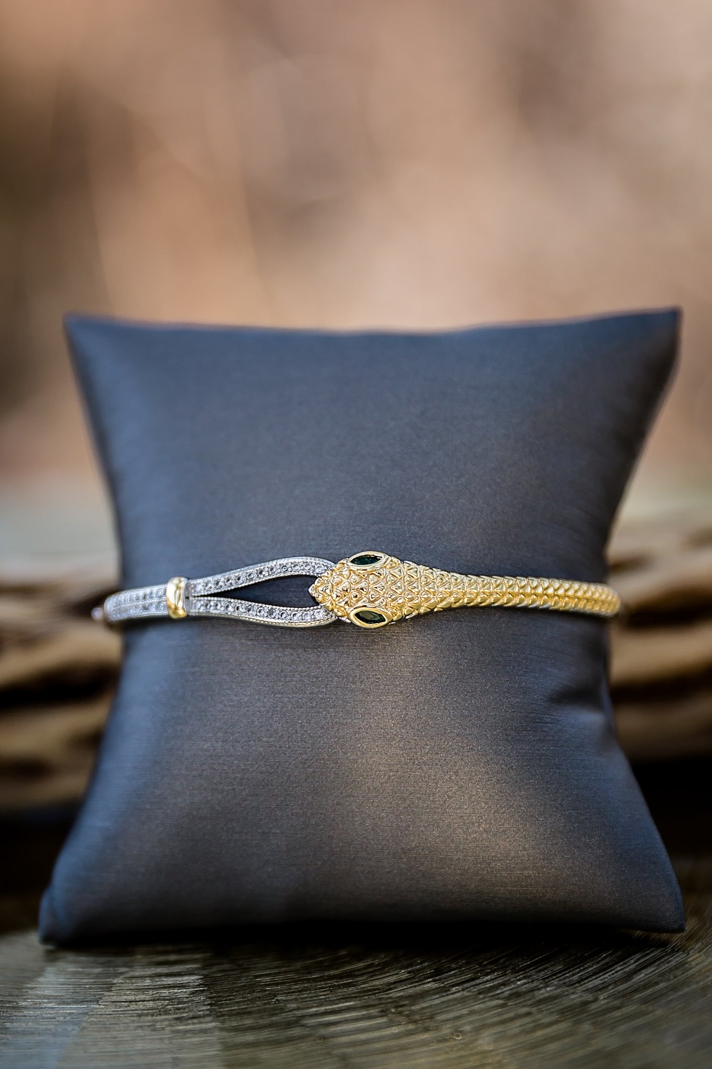 Wild Asp snake bangle displayed on pillow featuring 18kt gold plating, green stone eyes, and hinged closure