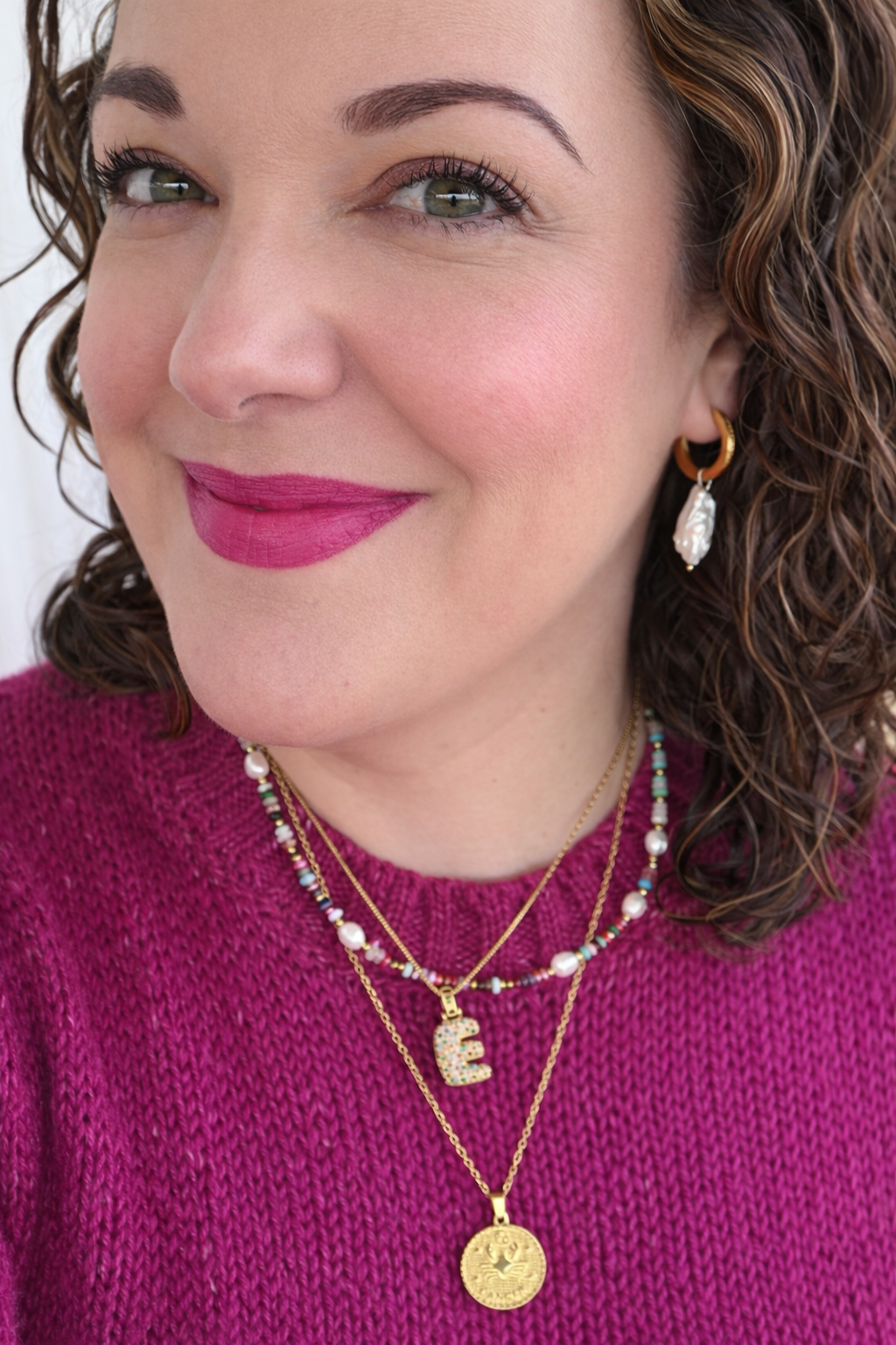 Woman wearing layered necklaces and earrings with a pink top against a white background