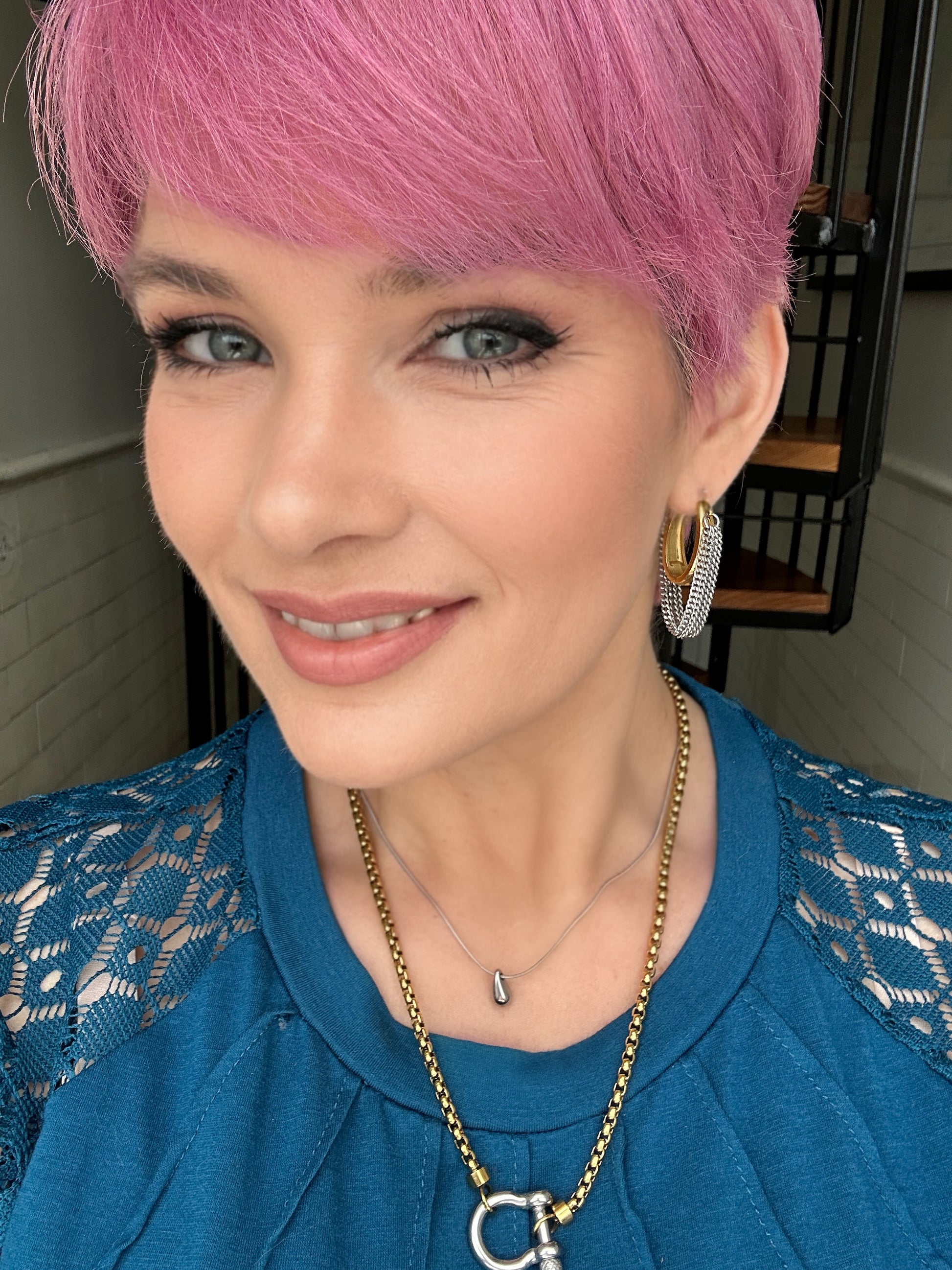 Woman with pink hair wearing a blue top and gold jewelry indoors.