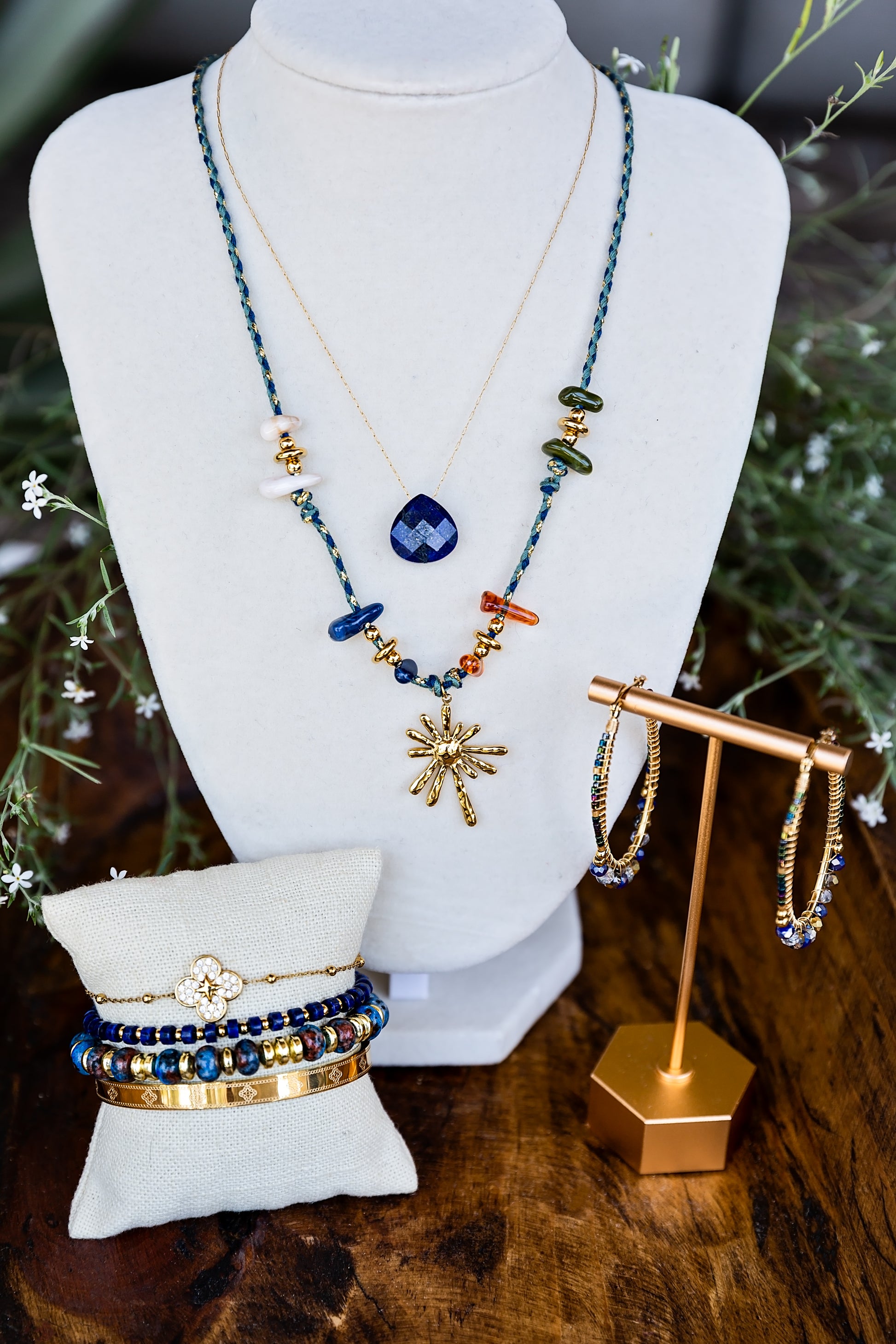 A jewelry display featuring a layered necklace set on a white mannequin bust, with matching beaded bracelets on a small pillow and gold hoop earrings on a stand, arranged on a wooden surface with greenery in the background.