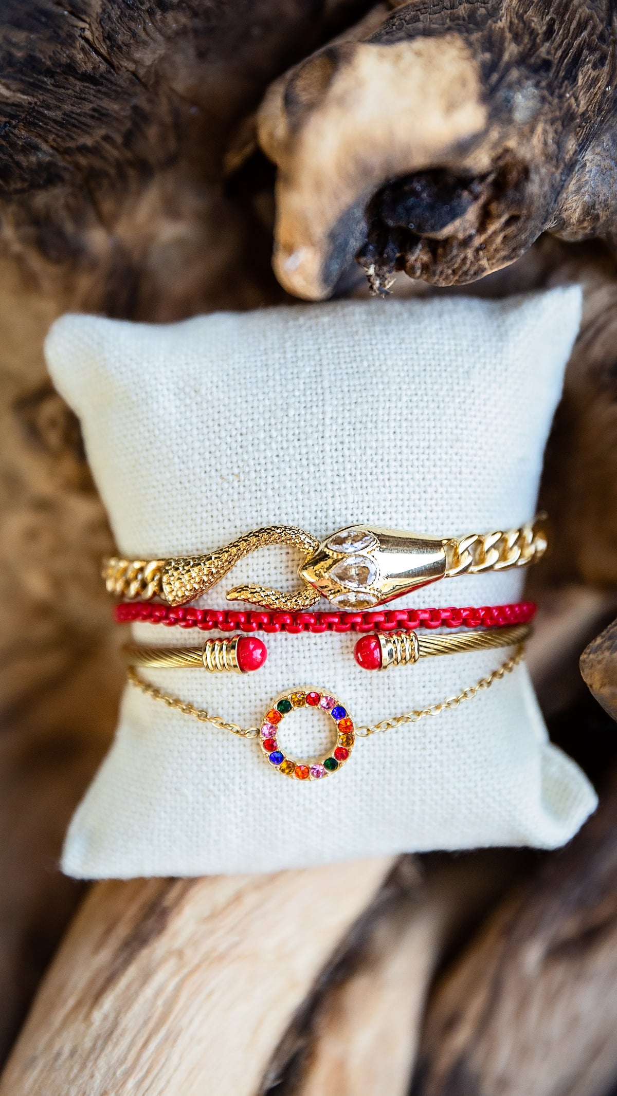 A stack of four gold and red bracelets displayed on a white fabric pillow, set against a rustic wood background. The bracelets include a gold snake charm, a red beaded bracelet, a gold bangle with red beads, and a gold chain with a colorful gemstone ring charm.