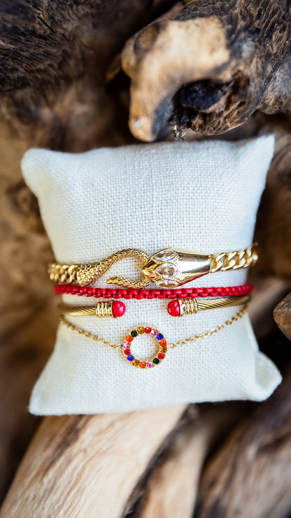 A stack of four gold and red bracelets displayed on a white fabric pillow, set against a rustic wood background. The bracelets include a gold snake charm, a red beaded bracelet, a gold bangle with red beads, and a gold chain with a colorful gemstone ring charm.