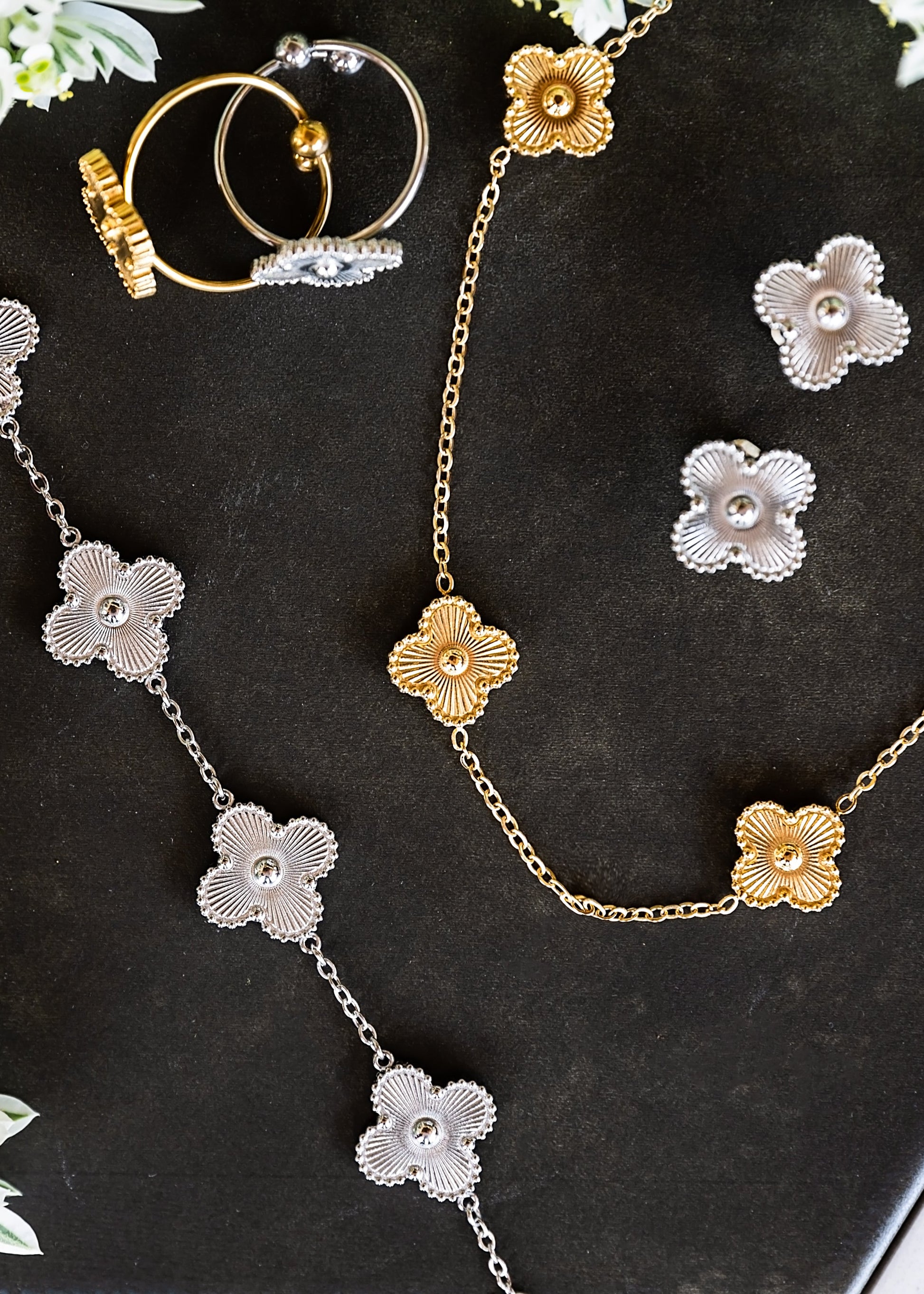 A jewelry flat-lay featuring a gold and silver floral chain necklace, matching flower earrings, and a gold ring set on a black tray with decorative green foliage.