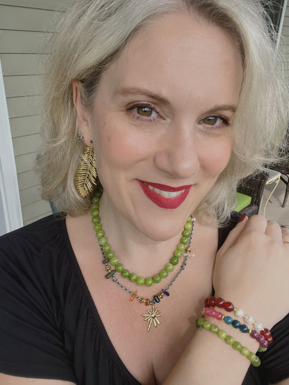 A close-up lifestyle shot of a woman with blonde hair wearing a green beaded necklace with a gold sunburst pendant, matching green beaded bracelets, and gold leaf earrings.