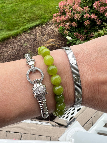 Three bracelets worn on a wrist outdoors: a silver-toned beaded bracelet with green beads, a silver-toned textured metal bracelet, and a silver-toned rope bracelet with a rhinestone ring and charm. Background shows grass, flowers, and patio stones.