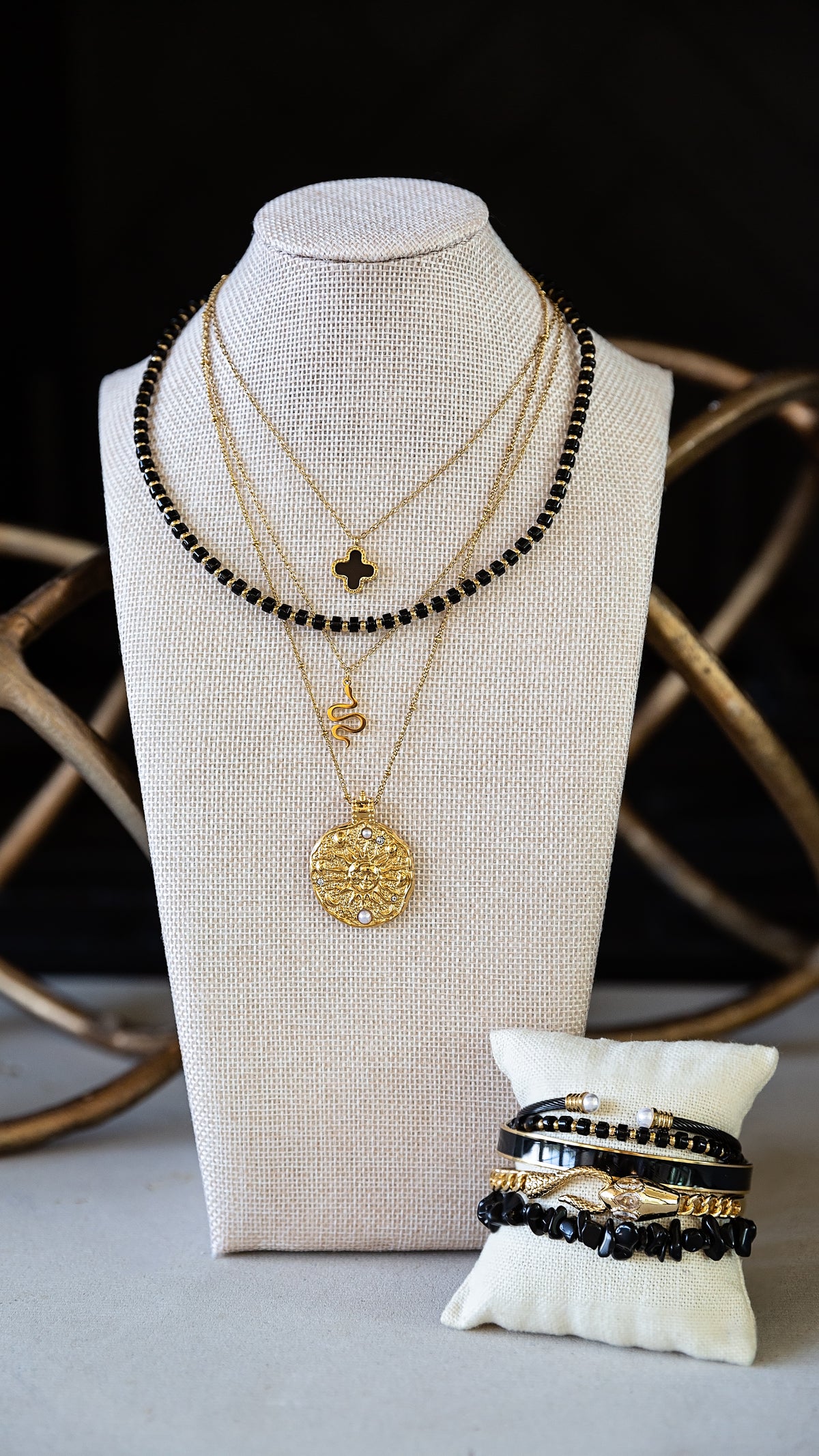 Layered gold and black beaded jewelry displayed on a cream fabric mannequin bust, with a small white pillow holding matching bracelets in front against a dark background with decorative gold wire elements.