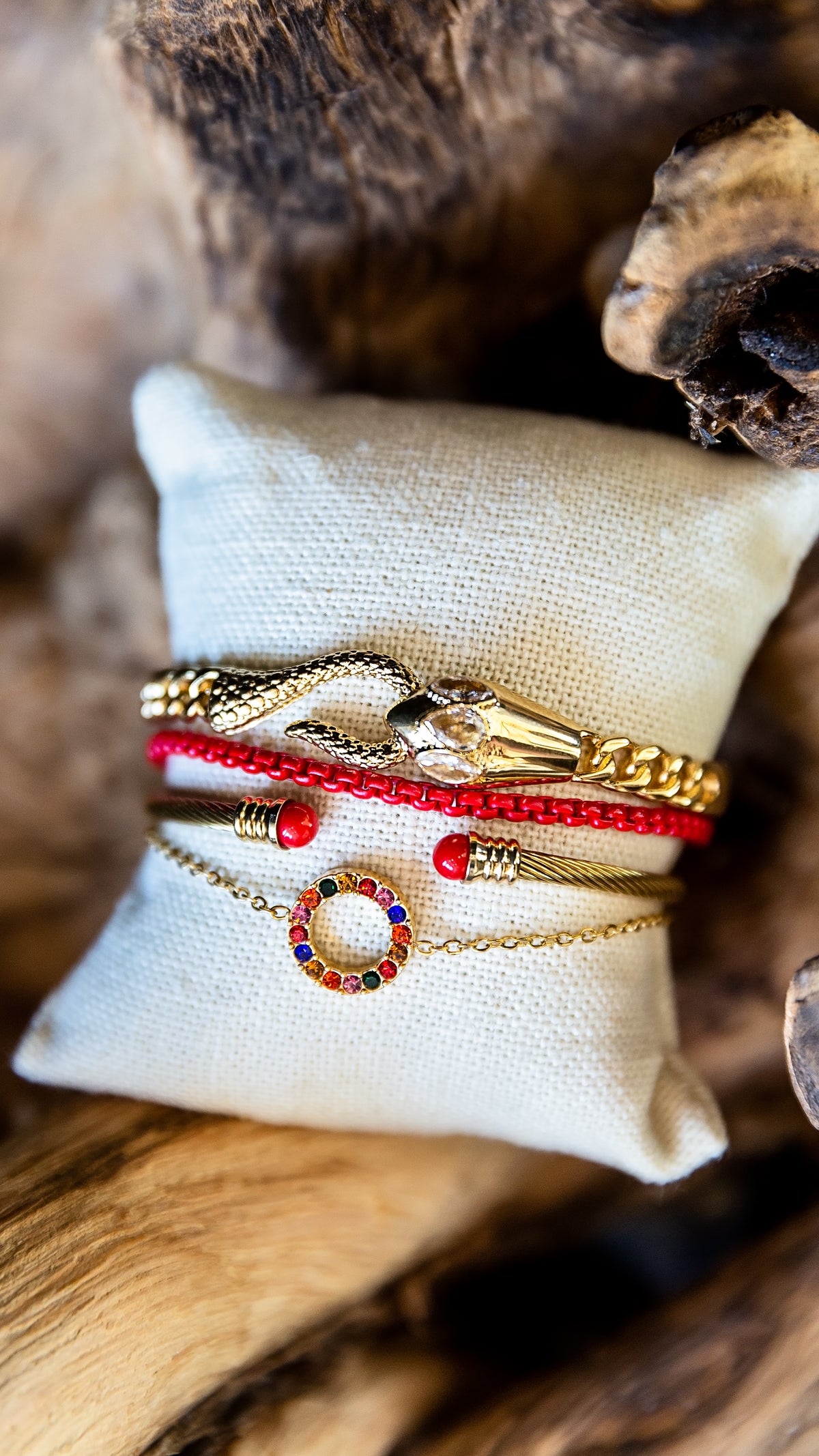 A set of four delicate bracelets displayed on a small beige fabric cushion, set against a rustic wood background. The bracelets include a gold-plated snake charm, a red beaded cord bracelet, a gold chain with red beads, and a gold chain featuring a colorful circular charm.