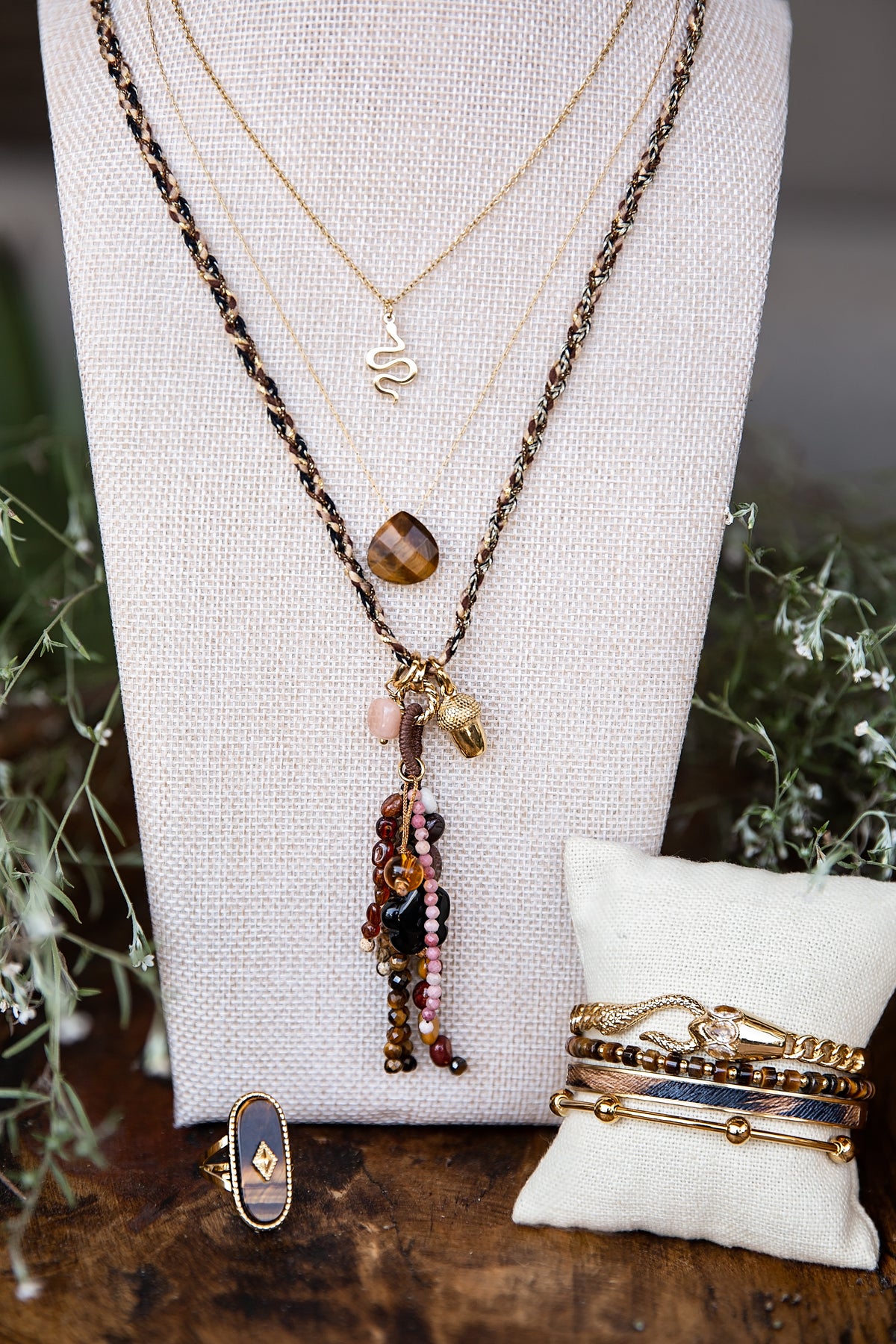 Layered Terra inspired  jewelry displayed on a cream fabric bust, featuring a long beaded necklace with a tiger's eye pendant, a gold snake necklace, and stacked gold and beaded bracelets on a small cream pillow, with a ring on a wooden surface.