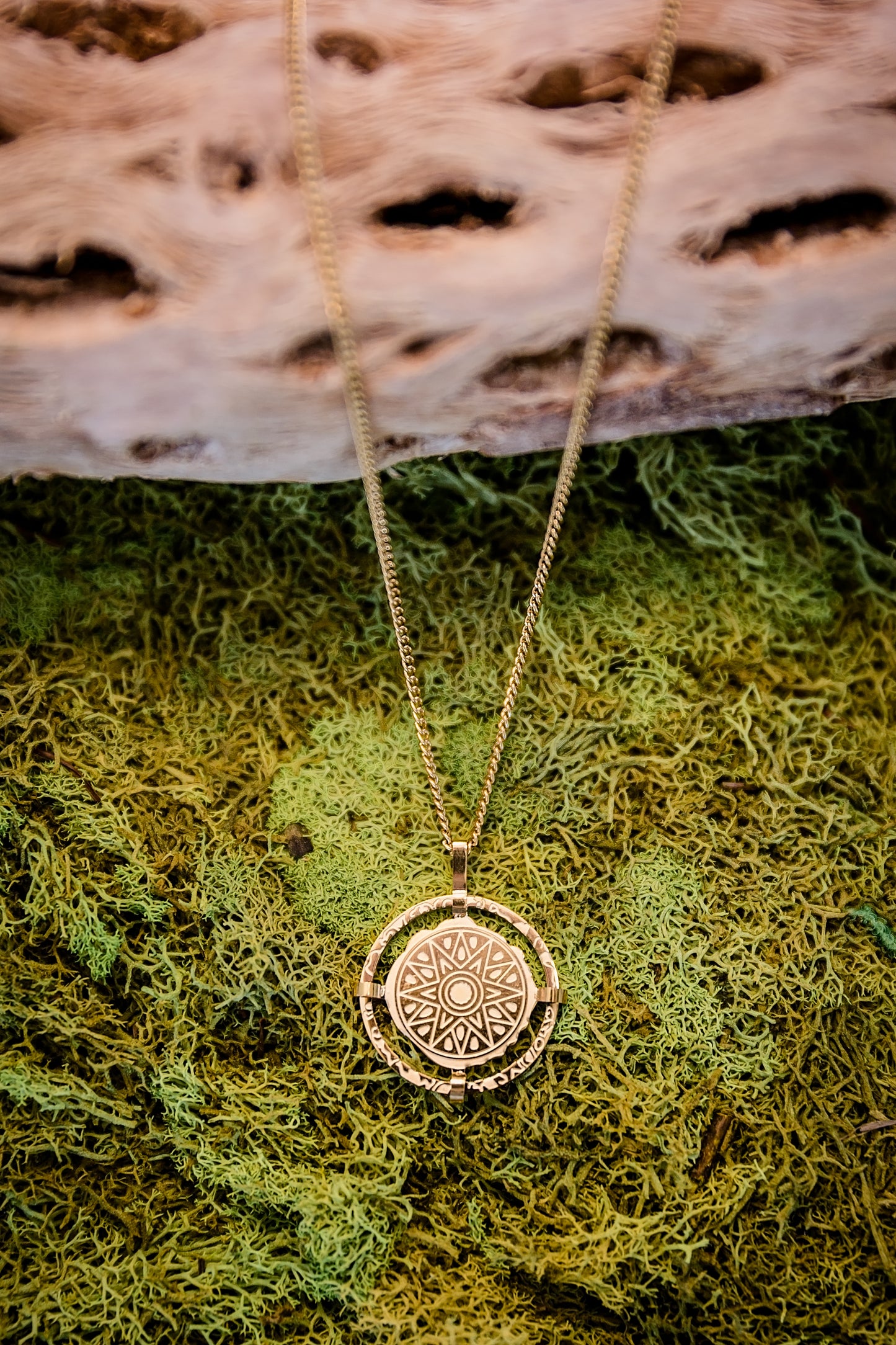 Gold compass pendant necklace with detailed etched design displayed on natural moss background