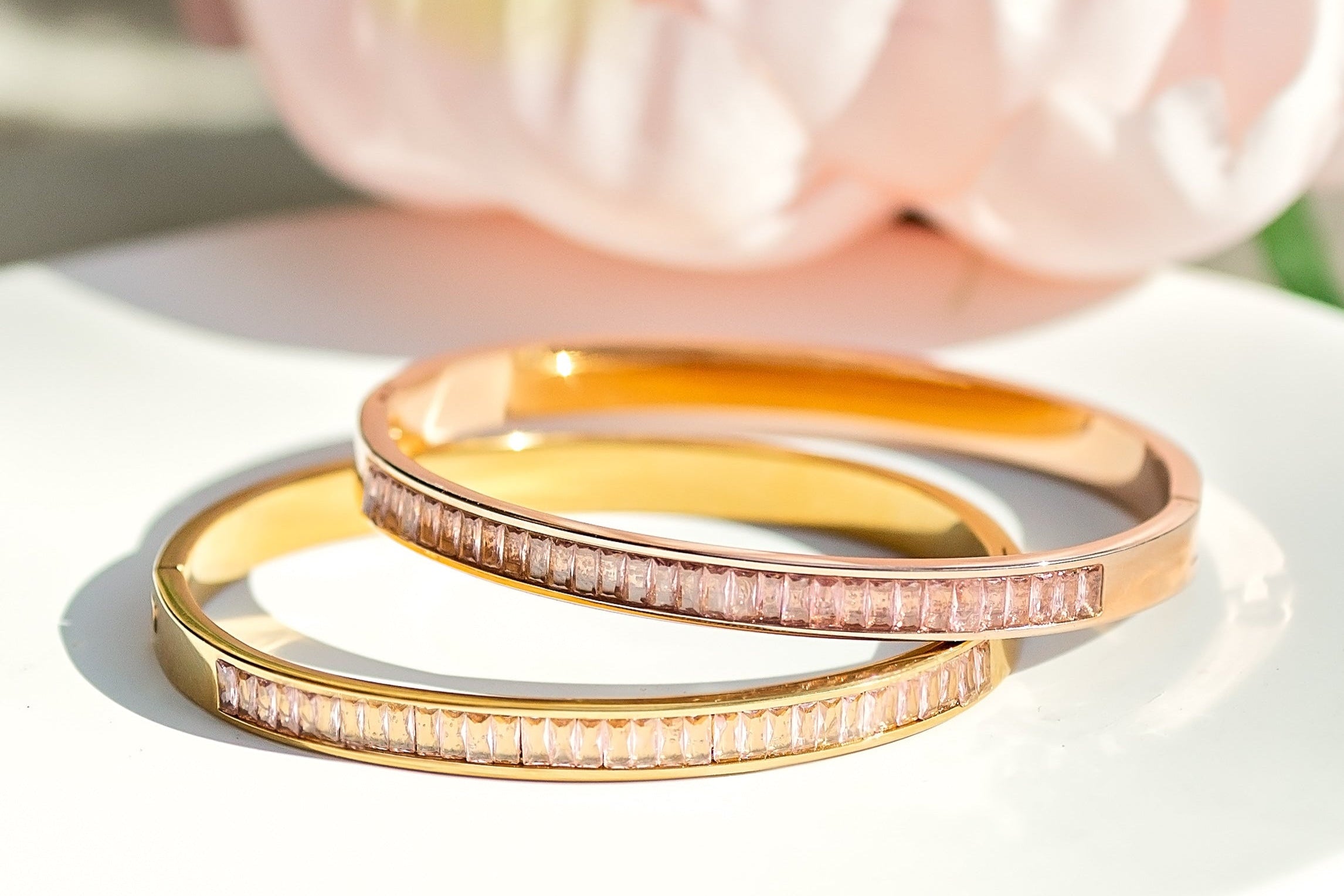 Two gold bracelets on a white plate with a pink flower in the background