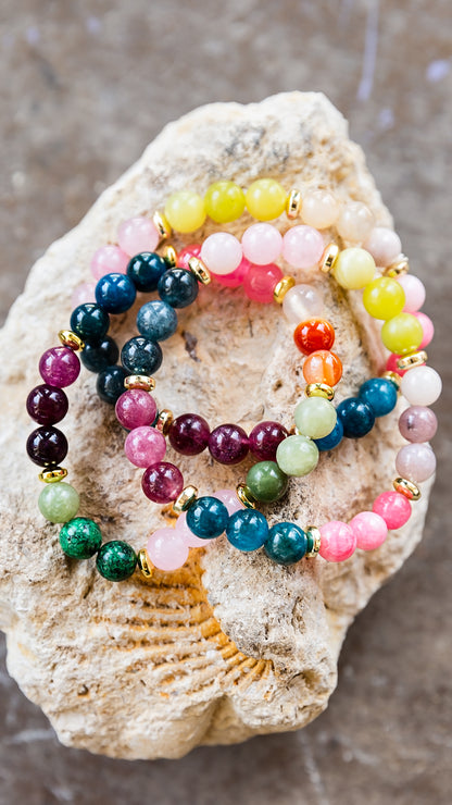 Three beaded stretch bracelets with gold spacer rings, featuring colorful gemstone beads, displayed on a textured beige stone background.