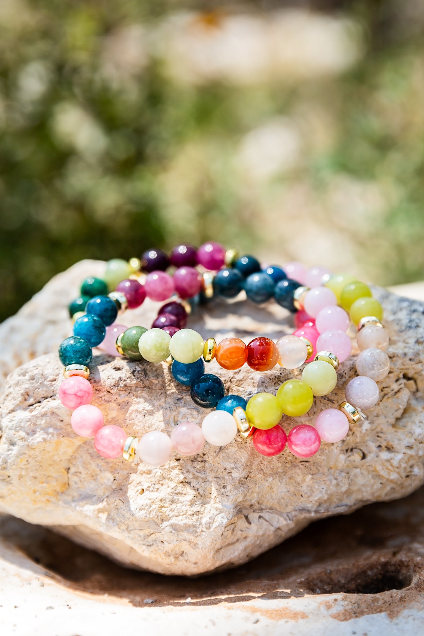 Three colorful beaded stretch bracelets with gold spacer rings, displayed on a light-colored stone with a blurred outdoor background.