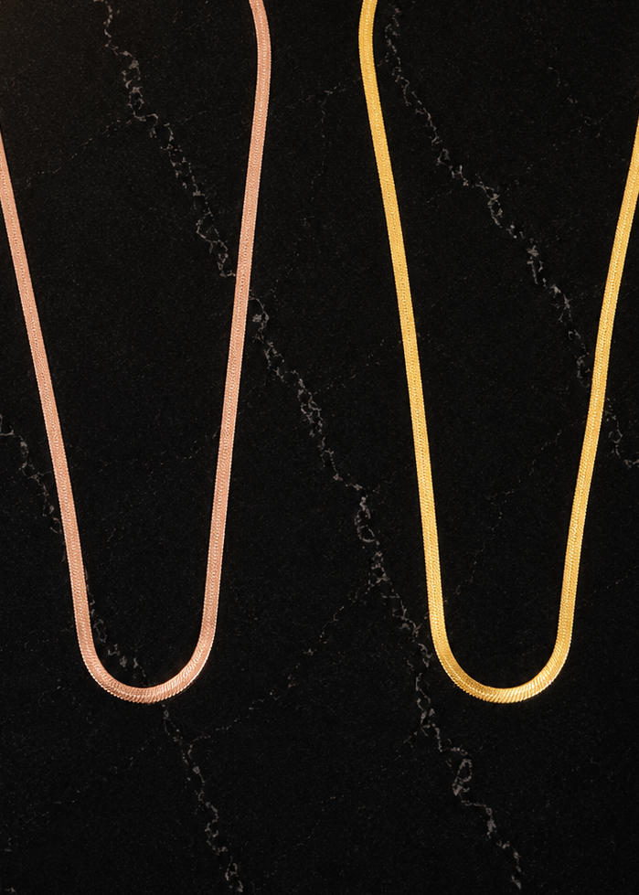Two slim chain necklaces displayed side by side on a dark marble-patterned background. The left necklace has a rose gold finish, and the right necklace has a yellow gold finish.