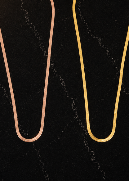 Two slim chain necklaces displayed side by side on a dark marble-patterned background. The left necklace has a rose gold finish, and the right necklace has a yellow gold finish.