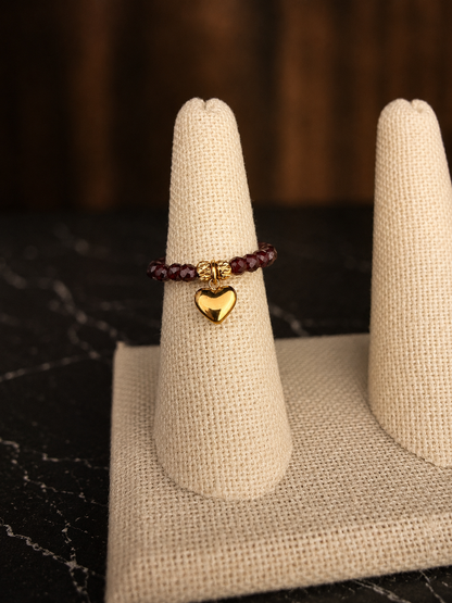 Claire Stretch Ring featuring garnet faceted gemstone beads and a gold heart charm displayed on a ring cone.