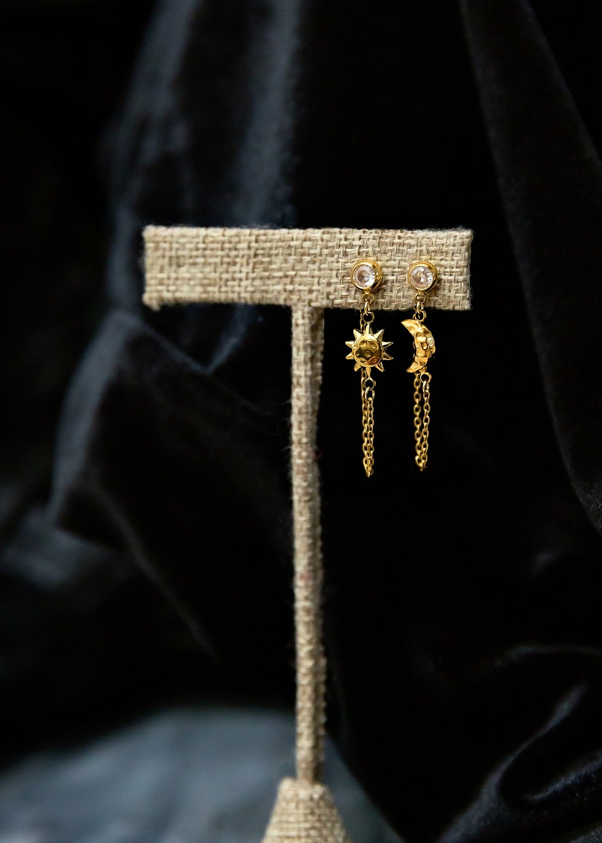 A pair of gold dangling earrings displayed on a woven fabric stand against a dark draped velvet background.