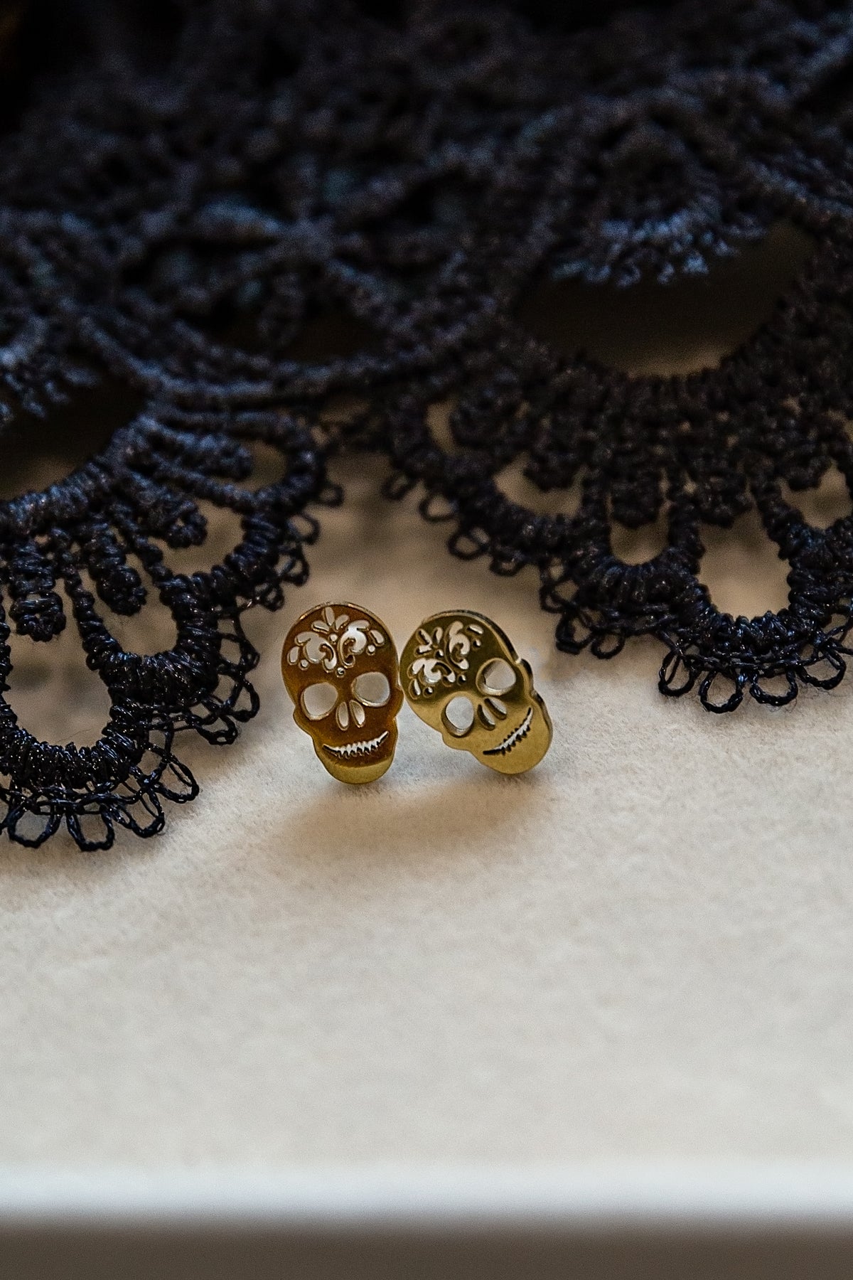Two small gold skull stud earrings resting on a cream-colored surface with a black lace prop in the background.