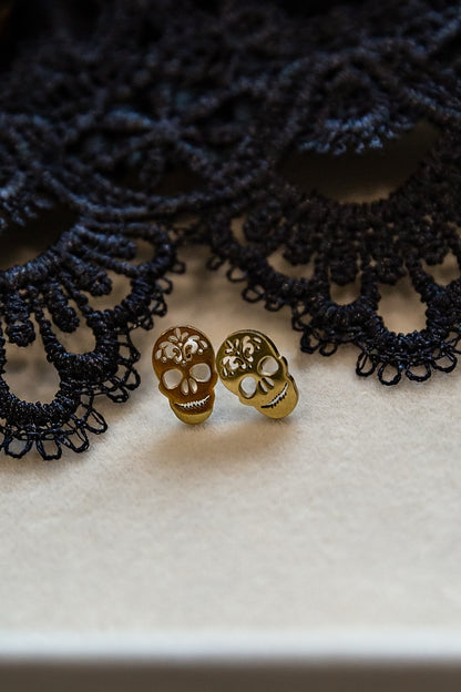 Two small gold skull stud earrings resting on a cream-colored surface with a black lace prop in the background.