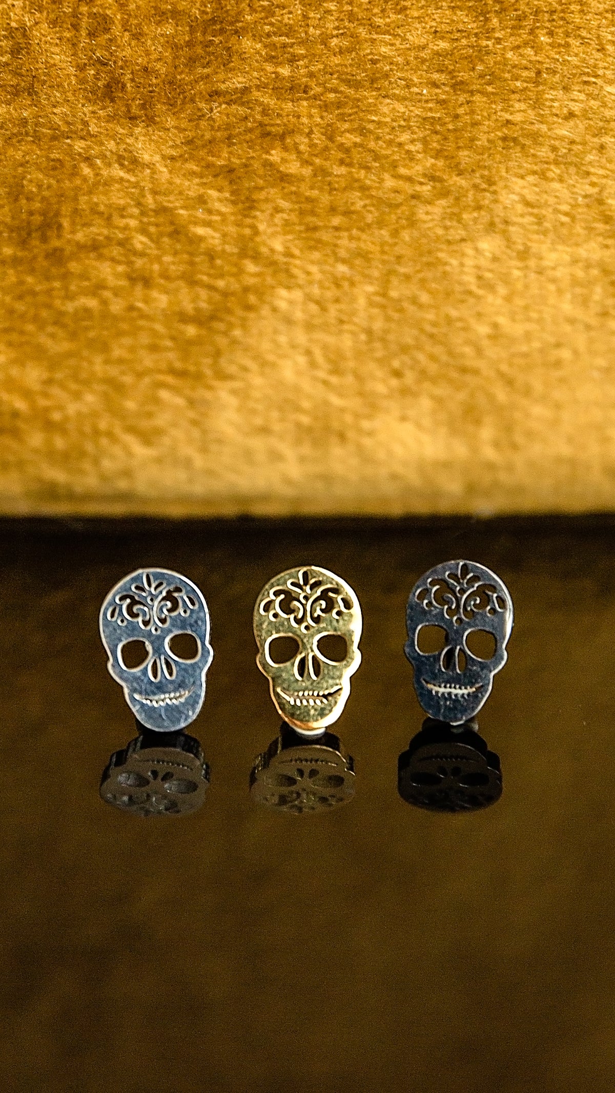 Three sugar skull stud earrings in silver, gold, and blue-gray finishes, arranged on a reflective dark surface against a textured gold background.