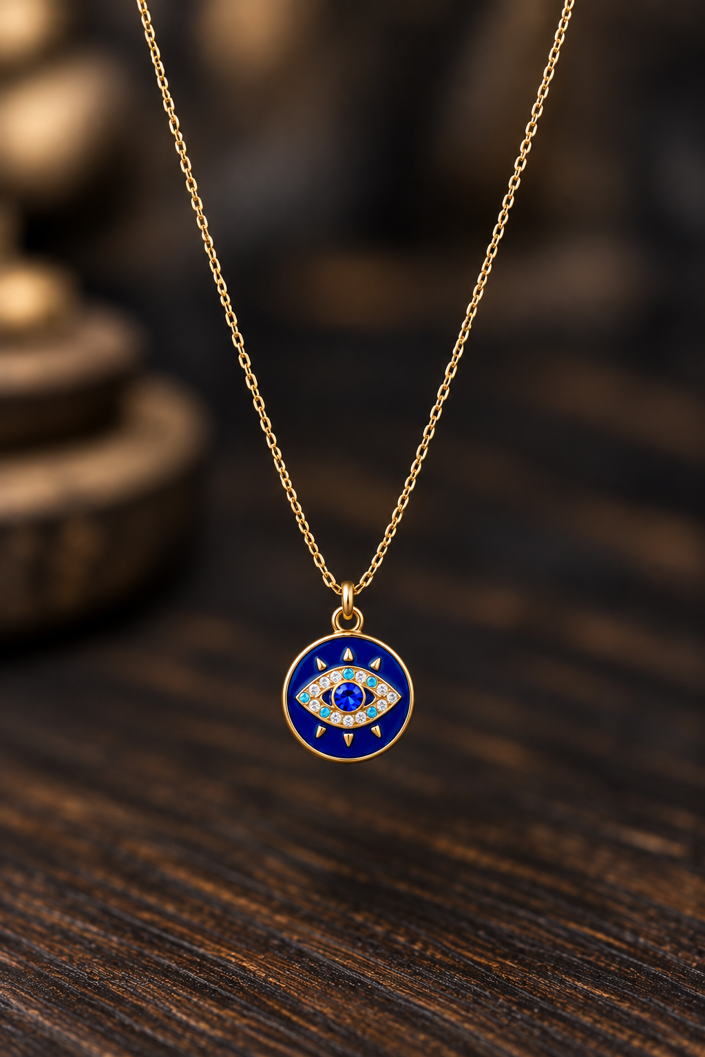 A gold chain necklace with a circular evil eye pendant featuring a deep blue enamel background and small white and red details, displayed on a dark wooden surface.