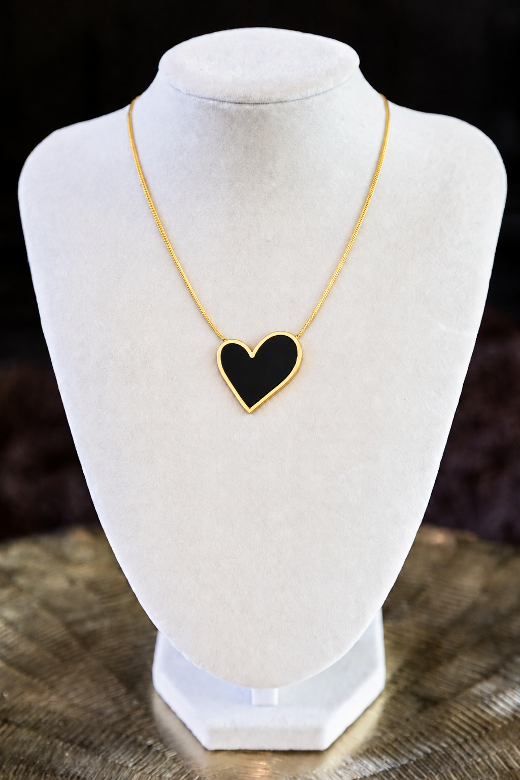 A gold chain necklace with a heart-shaped pendant featuring a black center and gold border, displayed on a white jewelry bust.