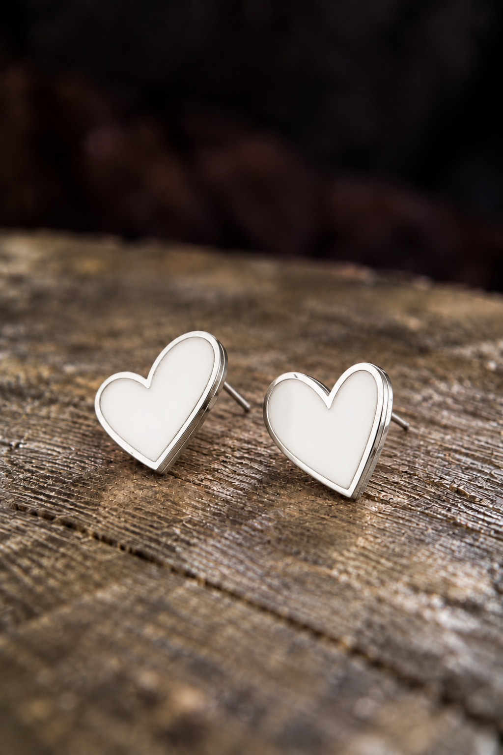 A pair of silver heart stud earrings with white enamel centers, resting on a rustic wooden surface with a dark, blurred background.