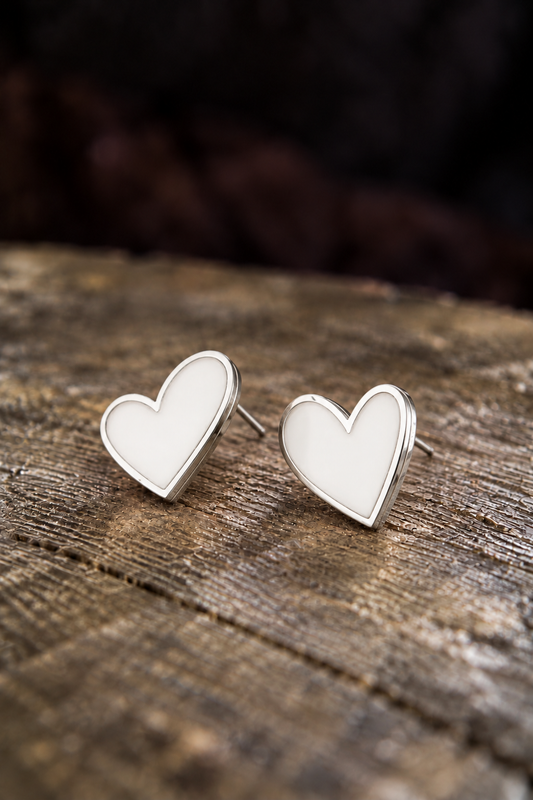A pair of silver heart stud earrings with white enamel centers, resting on a rustic wooden surface with a dark, blurred background.