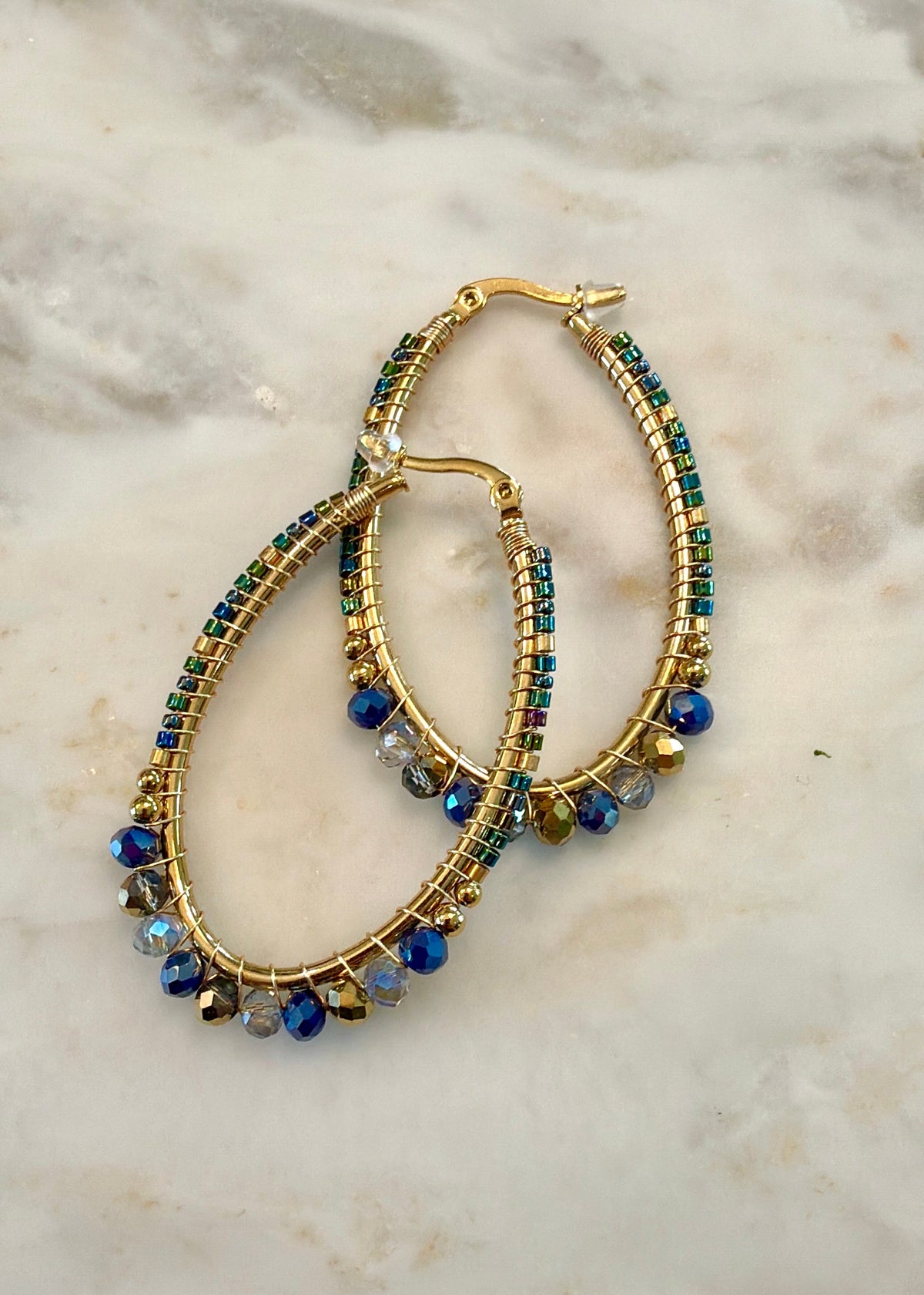 Gold hoop earrings with rows of green and blue beads and dangling blue beads, displayed on a light marble surface.