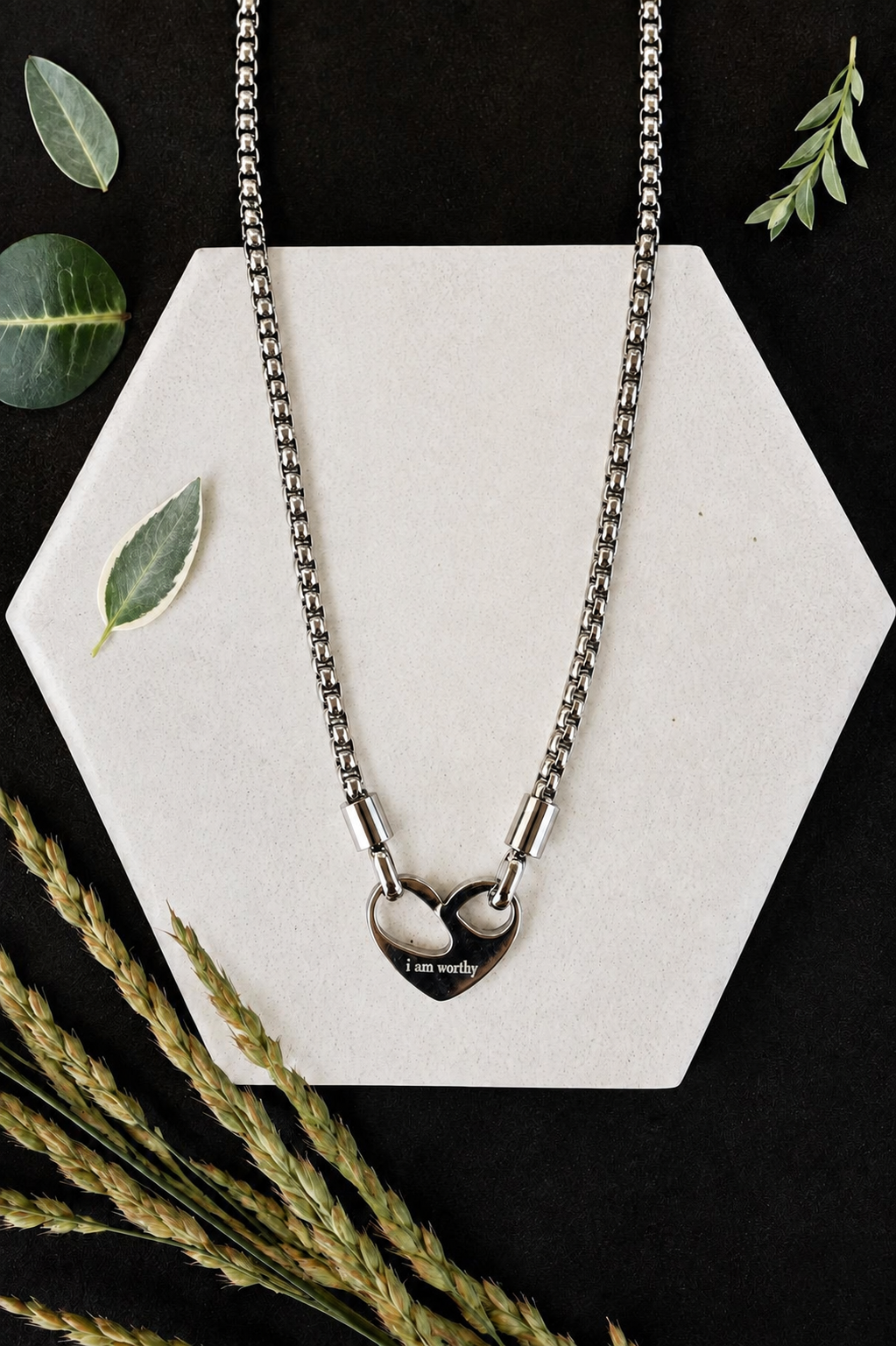 A silver-toned chain necklace with two interlocking heart pendants, one engraved with 'I am worthy', displayed on a white hexagonal card with decorative leaves and wheat stems on a black background.