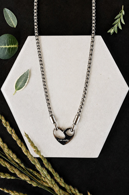 A silver-toned chain necklace with two interlocking heart pendants, one engraved with 'I am worthy', displayed on a white hexagonal card with decorative leaves and wheat stems on a black background.