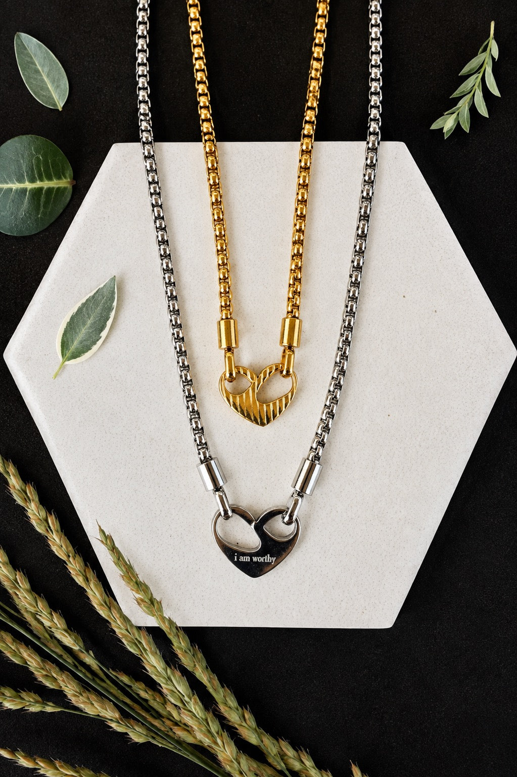 Two heart-themed necklaces displayed on a white hexagonal board against a black background, styled with dried wheat stalks and green leaves. One necklace is gold with a heart pendant, and the other is silver with a split heart pendant engraved with 'I am worthy'.