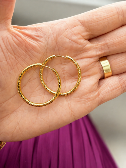 A pair of textured gold hoop earrings resting on an open hand, with a matching gold ring visible on the finger. The background is softly blurred, showing a gray wall and a purple garment.