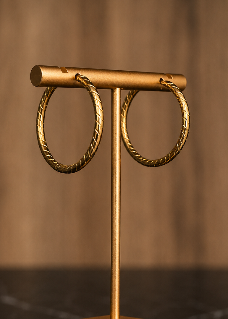 Gold-tone metal jewelry display stand holding a pair of large twisted hoop earrings, set on a dark marble surface against a warm blurred wood background.