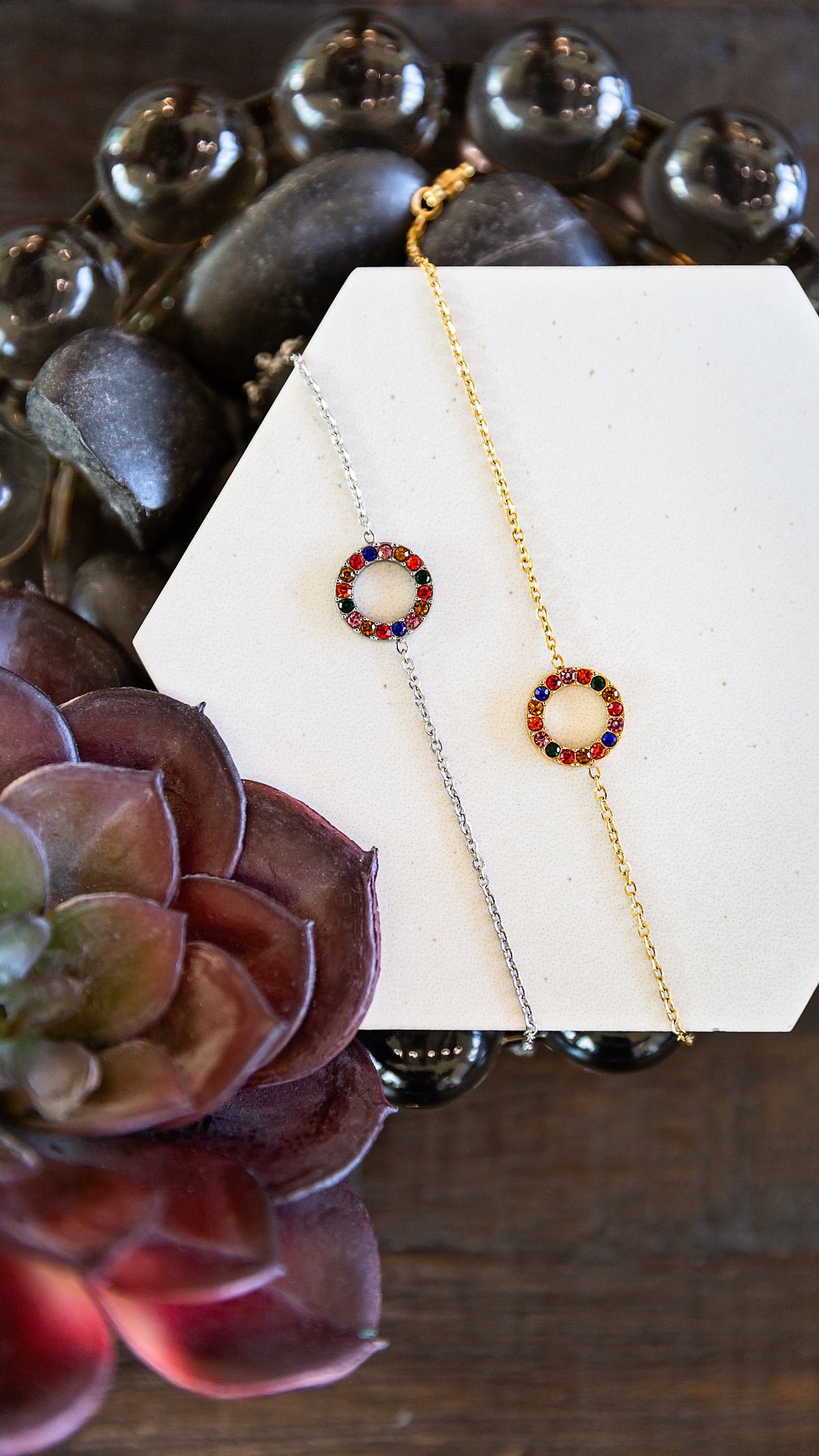 Two delicate chain bracelets displayed on a white geometric card, one gold and one silver, both featuring a small circular ring set with red, blue, and green stones. The card is styled on a dark wooden surface with dark decorative stones and a purple succulent in the foreground.