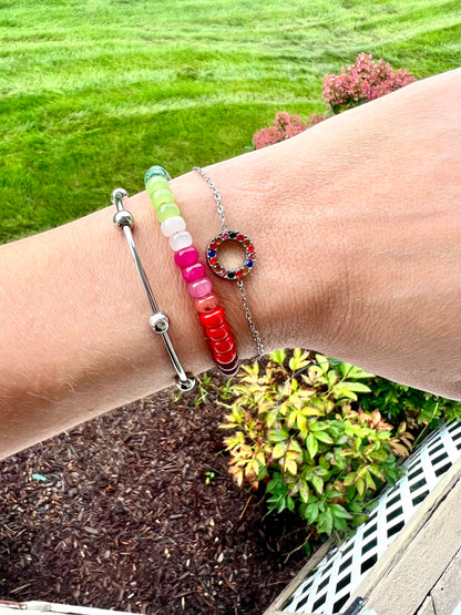 Three beaded bracelets worn on a person's wrist, with a green grass and garden bed in the background. The bracelets include a silver ball-beaded bracelet, a pastel rainbow bead bracelet, and a silver chain bracelet with a colorful circular bead charm.