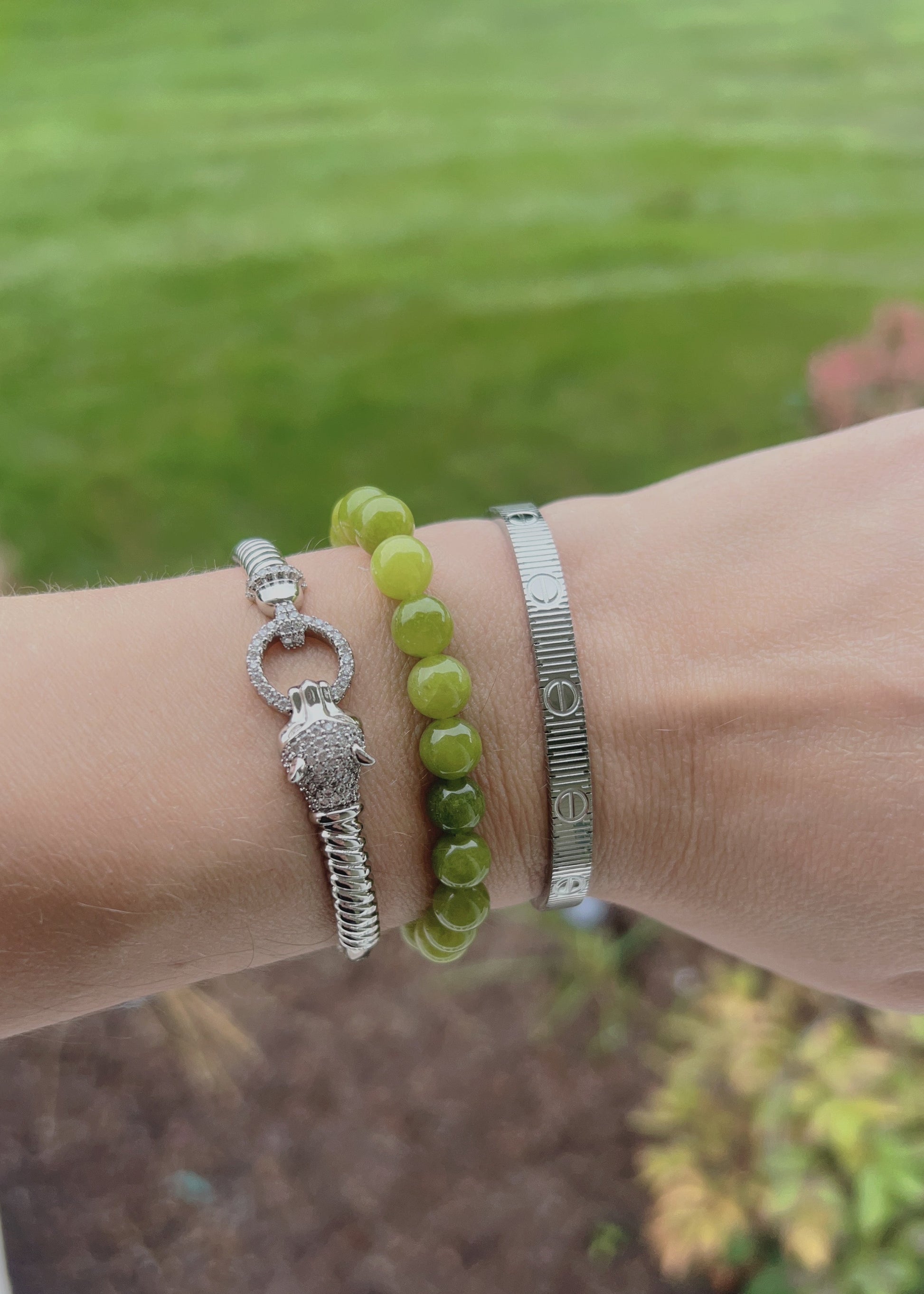 Three bracelets worn on a wrist: a silver turtle charm bracelet, a green beaded bracelet, and a textured silver cuff bracelet, with a blurred outdoor garden background.