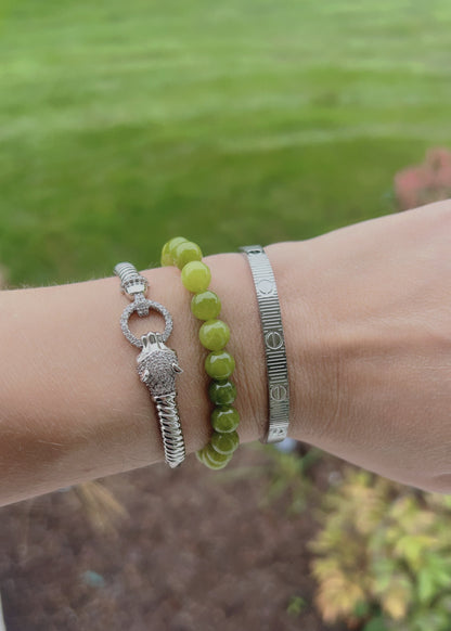 Three bracelets worn on a wrist: a silver turtle charm bracelet, a green beaded bracelet, and a textured silver cuff bracelet, with a blurred outdoor garden background.