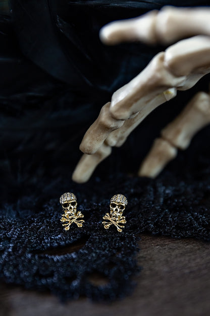 Small crystal-accented skull stud earrings by VEITA Jewelry photographed with dark textured fabric and natural decorative elements.