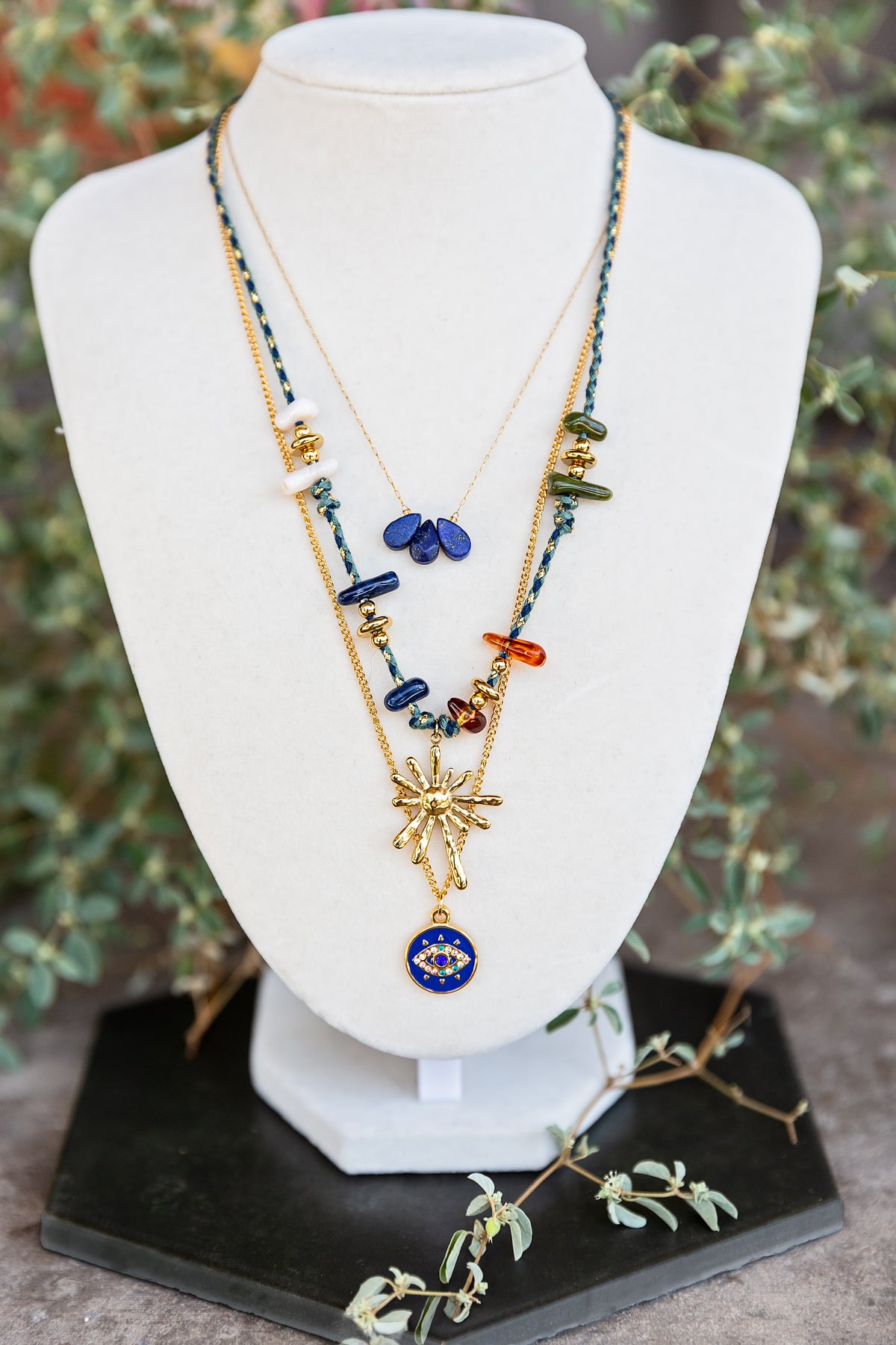 Trine Necklace styled with layered necklaces on a white jewelry display, featuring colorful genuine gemstone details.