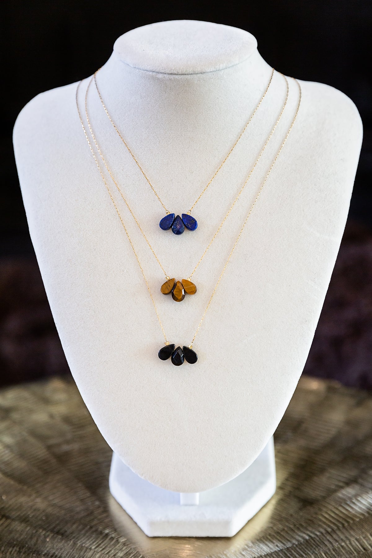 Trine Necklace with three layered gold chains and blue, amber and black gemstone drops displayed on a white jewelry bust.