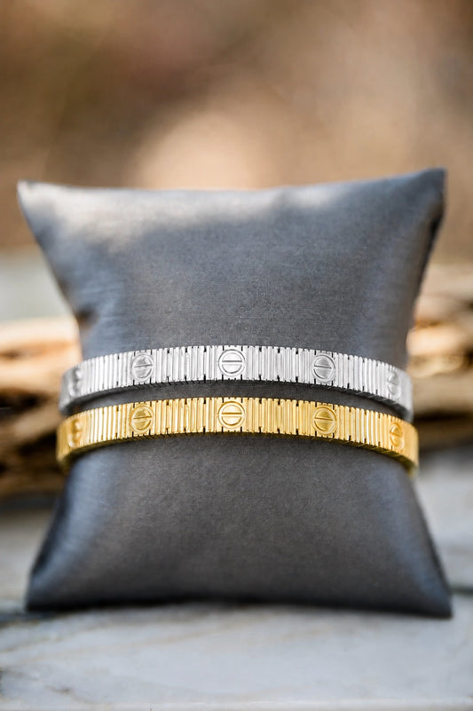 Two bangle bracelets, one silver and one gold, displayed on a dark gray fabric pillow. The bracelets feature a textured, ribbed design with engraved circular details.
