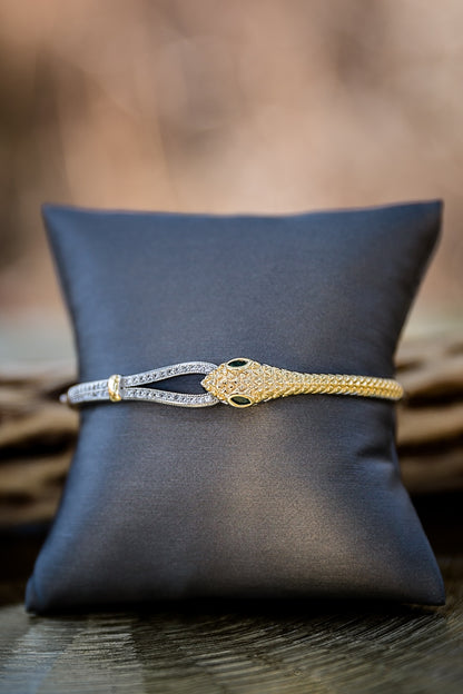 Wild Asp snake bangle displayed on pillow featuring 18kt gold plating, green stone eyes, and hinged closure